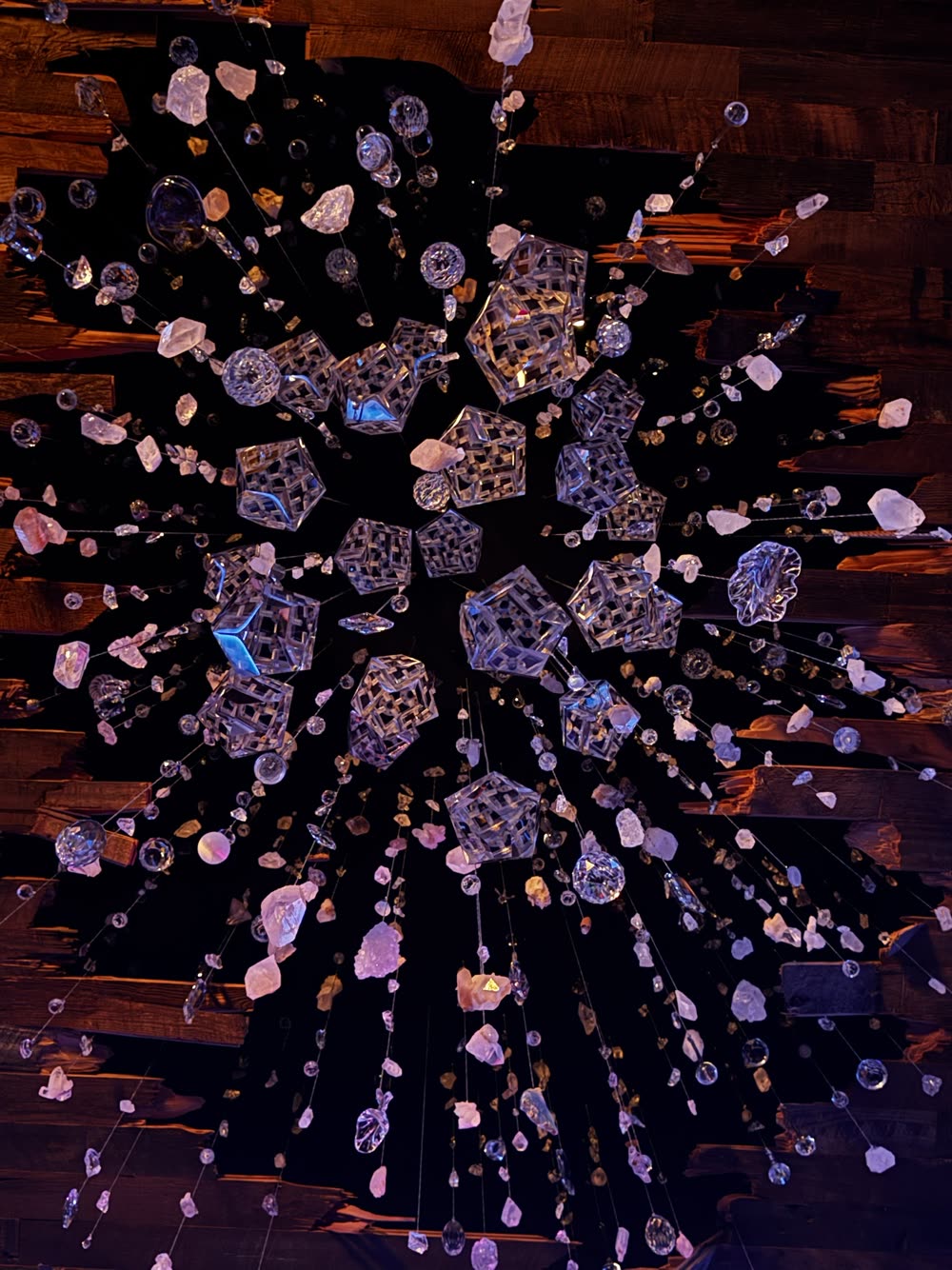 Directly beneath the installation: faceted crystal forms at the center with strands of rose and clear quartz radiating out toward the broken wood lath.
