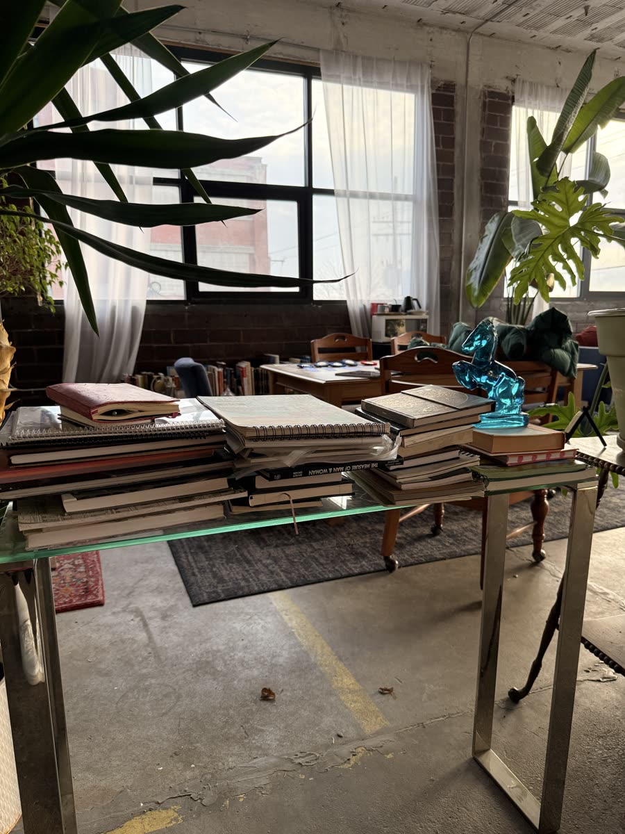 Stacks of sketchbooks, notebooks and reference books on a glass table in the private studio, with a blue glass horse on top.