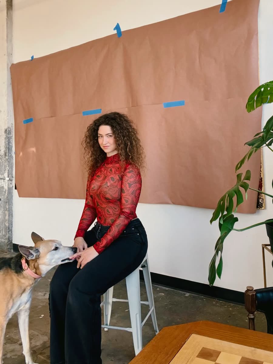Autumn Lundberg seated on a stool in her private studio, in front of a large canvas wrapped in brown paper, with her dog beside her.