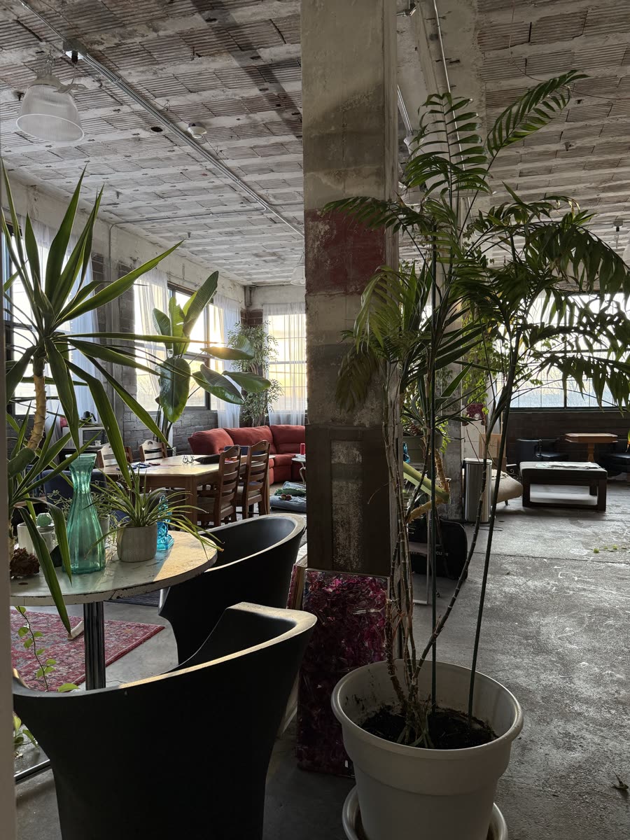 The private studio: an open warehouse loft with concrete columns, tall plants, a work table and a red sofa in evening light.